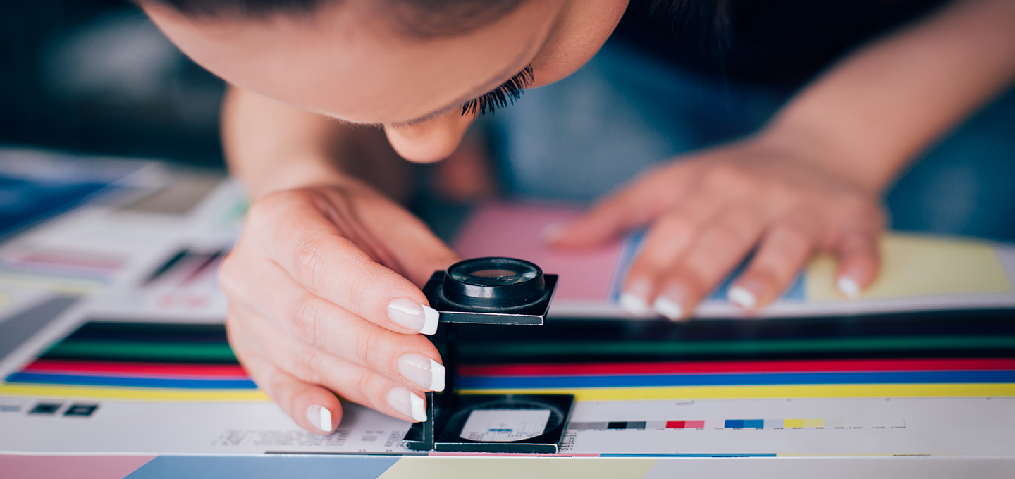 A woman is holding a magnifying device over a printed document which shows black and CYMK colours and specifications.