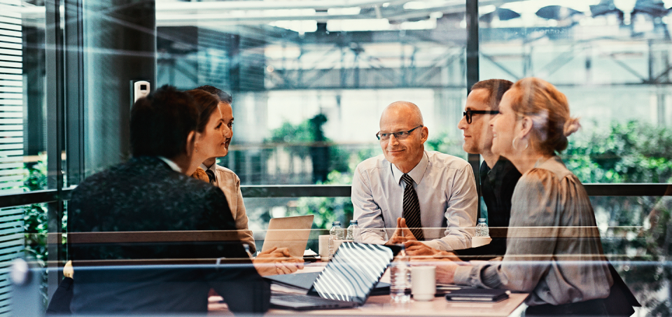 A meeting is being held in a modern meeting room with glass walls, looking out onto a clean office space. A group of colleagues are sitting around a desk with their papers and laptops in front of them. They are engaged in conversation and smiling at one another.