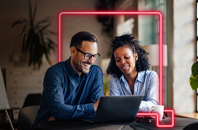 A man and woman smile while looking at a laptop in a warmly lit office with a whiteboard. The scene conveys collaboration and positivity.