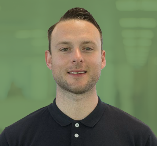 Photograph of a man with short hair wearing a black polo shirt, standing in a hallway with blurred lights and walls in the background. The focus is on the man, positioned on the left side, with a greenish tint covering the right side of the frame.