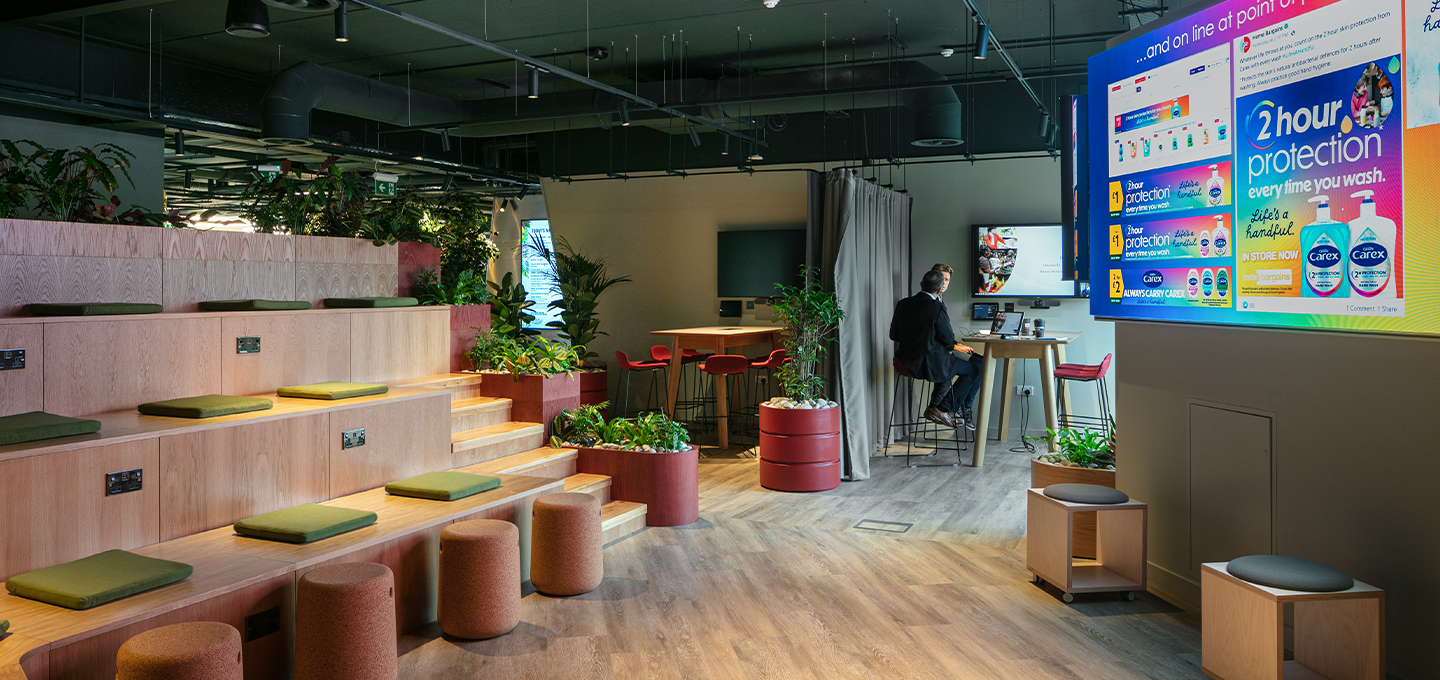 Modern collaborative workspace with tiered seating, plants, wooden furnishings, and a digital information screen in an open office environment.