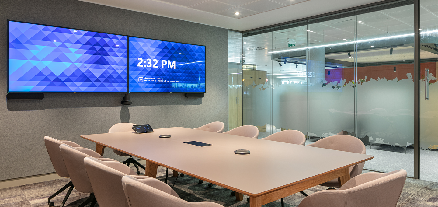 Modern meeting room with a large conference table, upholstered chairs, dual wall‑mounted screens, and glass walls in a contemporary office setting.