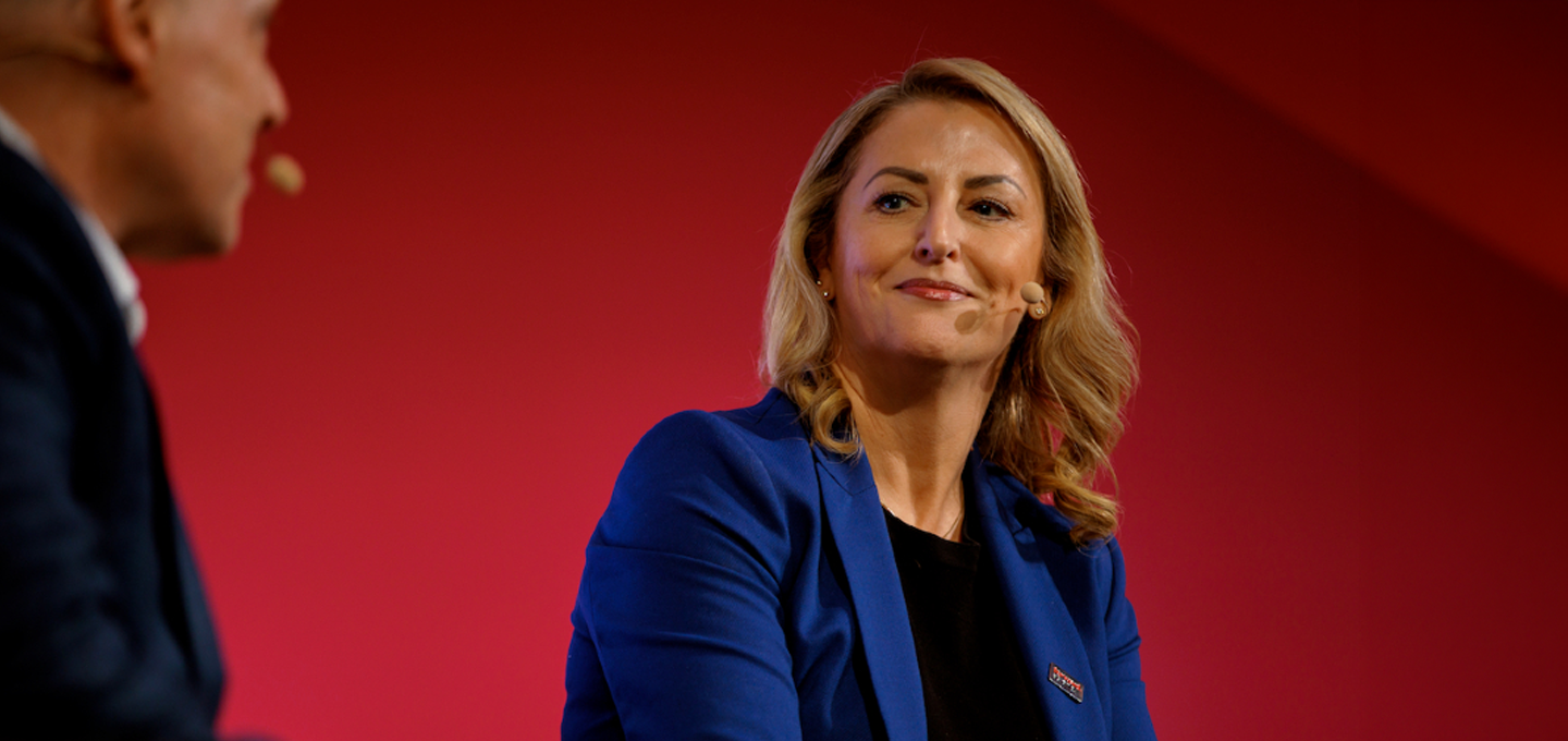 A woman in a blue blazer speaks with a man on stage against a red background. She appears confident and engaged, with a slight smile.