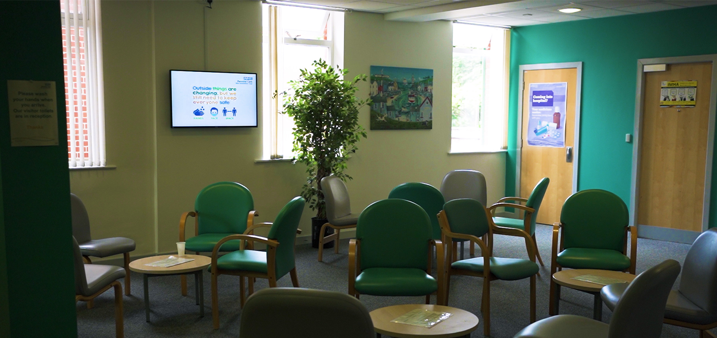 Healthcare waiting room with green chairs and digital information screens displaying patient messages.