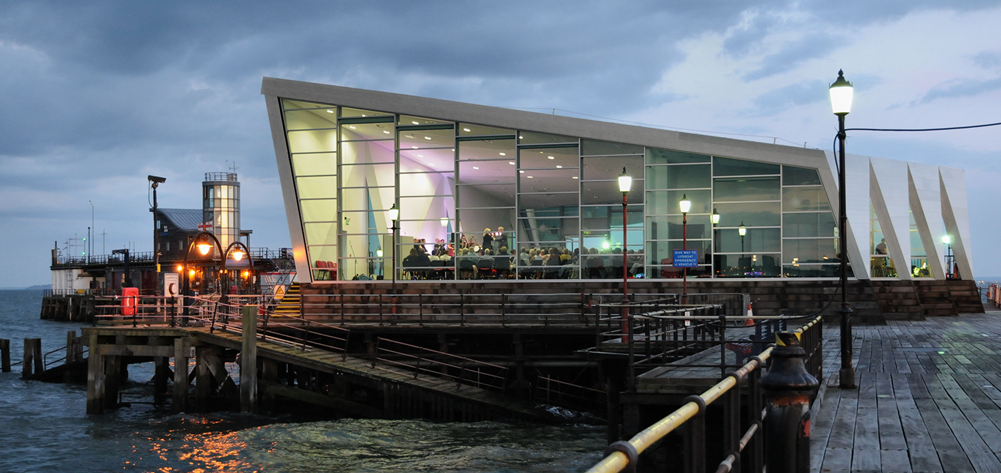 A twilight photograph captures the Southend Pier Head complex, showcasing the dramatic, modern, glass-fronted Southend Pier Cultural Centre (or Pier Head building) illuminated from within, standing over the water at the end of a wooden pier structure, with the older pier infrastructure and dark, cloudy sky framing the scene.