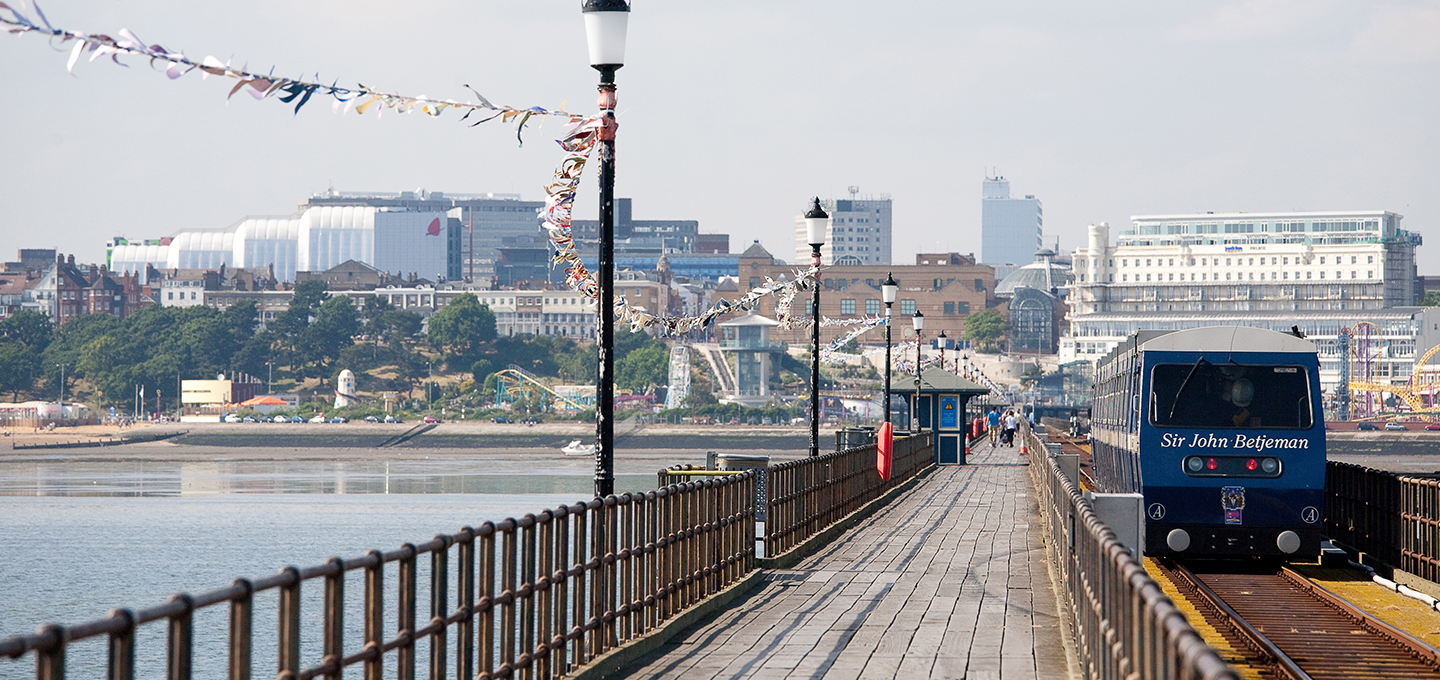 A sunny view along Southend Pier, Essex, showing the historic track, decorative bunting, and the Sir John Betjeman railway train against the background of the vibrant coastal town.