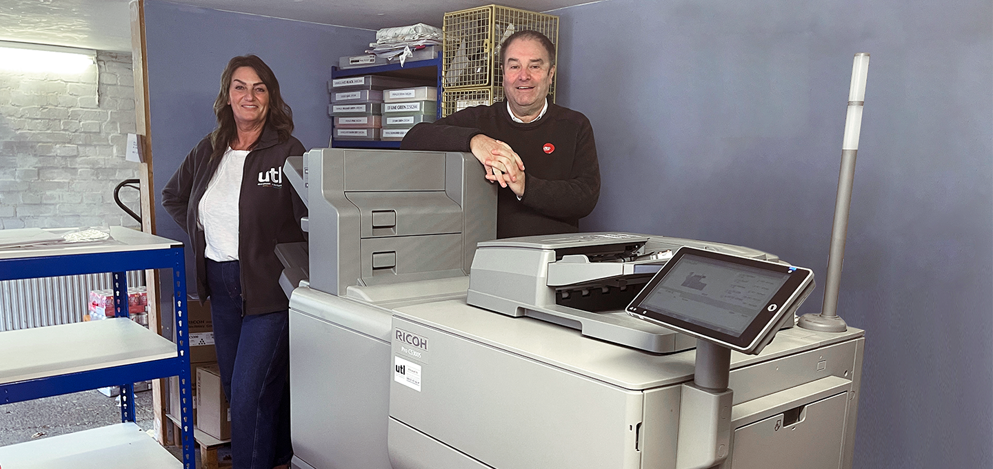 Two people standing beside a Ricoh industrial printer in a workspace with shelving and storage units.