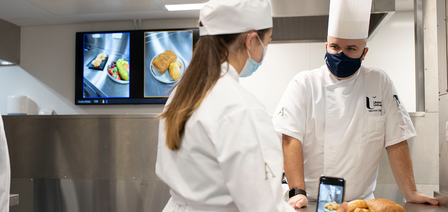 Photograph of two chefs in a commercial kitchen, one holding a smartphone displaying a photo of bread rolls, while the other listens attentively. Digital screens in the background show close-up images of plated food items, indicating a focus on food presentation or quality control.