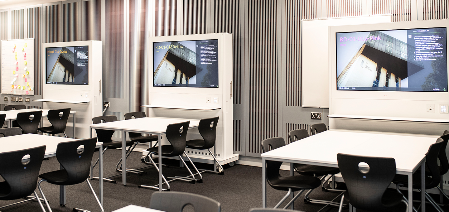 Photograph of a modern classroom featuring multiple white tables and black chairs arranged in rows, with three large digital screens mounted on white stands displaying the same presentation slide. The room has a clean, organized layout with vertical striped wall panels and a focus on technology integration for interactive learning.